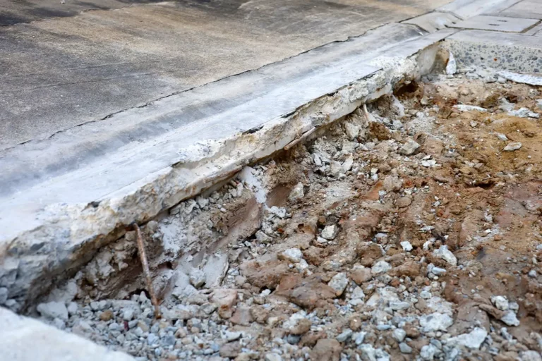 Durch Betonsägearbeiten beschädigter Gehweg mit freiliegendem Erdreich neben einer asphaltierten Straße.