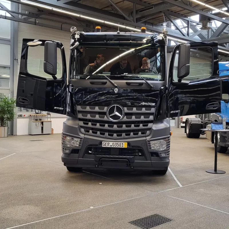 Ein schwarzer Mercedes-Lkw in einem Innenraum, beide Vordertüren sind geöffnet, im Fahrerhaus sitzen Personen.