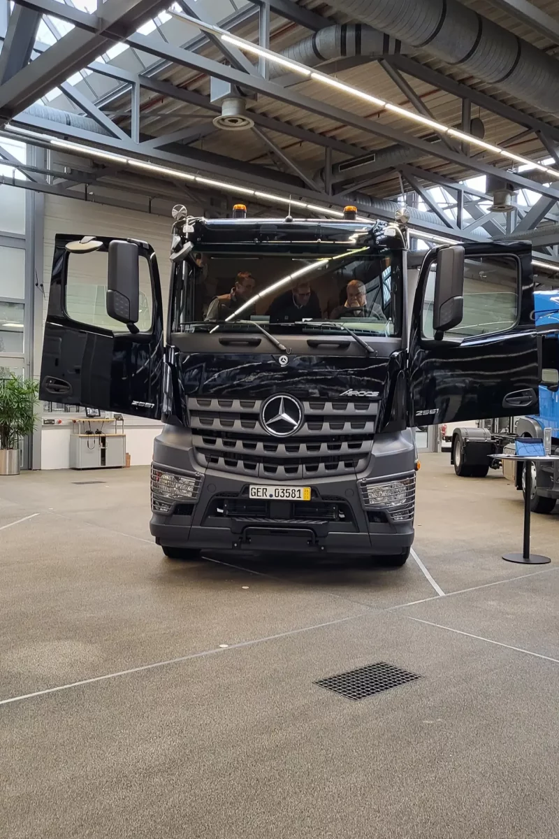 Ein schwarzer Mercedes-Lkw in einem Innenraum, beide Vordertüren sind geöffnet, im Fahrerhaus sitzen Personen.