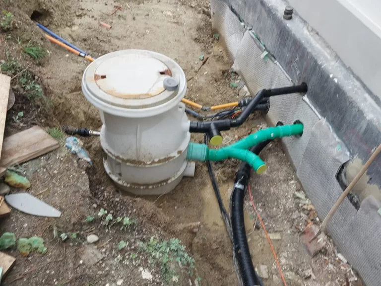 Ein weißer Außenablauf mit Entwässerungselementen und Rohren im Erdreich in der Nähe eines Gebäudes.