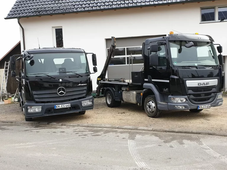 Vor einem weißen Gebäude mit einem Garagentor standen zwei schwarze Lastwagen.