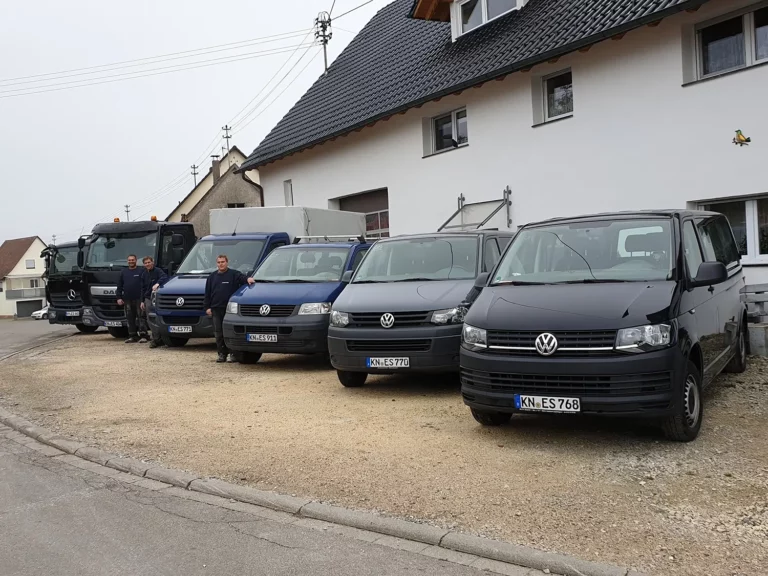 Vor einem weißen Haus stehen fünf Transporter und drei Personen.