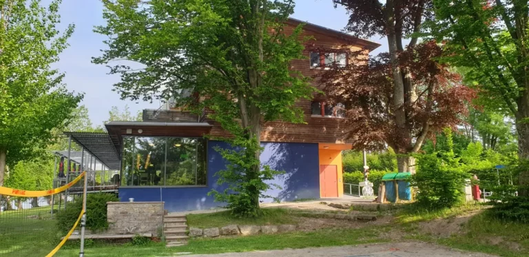 Ein modernes Haus mit Anbauten, blauen und orangefarbenen Wänden, umgeben von viel Grün.