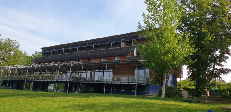 Modernes dreistöckiges Glasgebäude mit Anbauten und Bäumen davor.