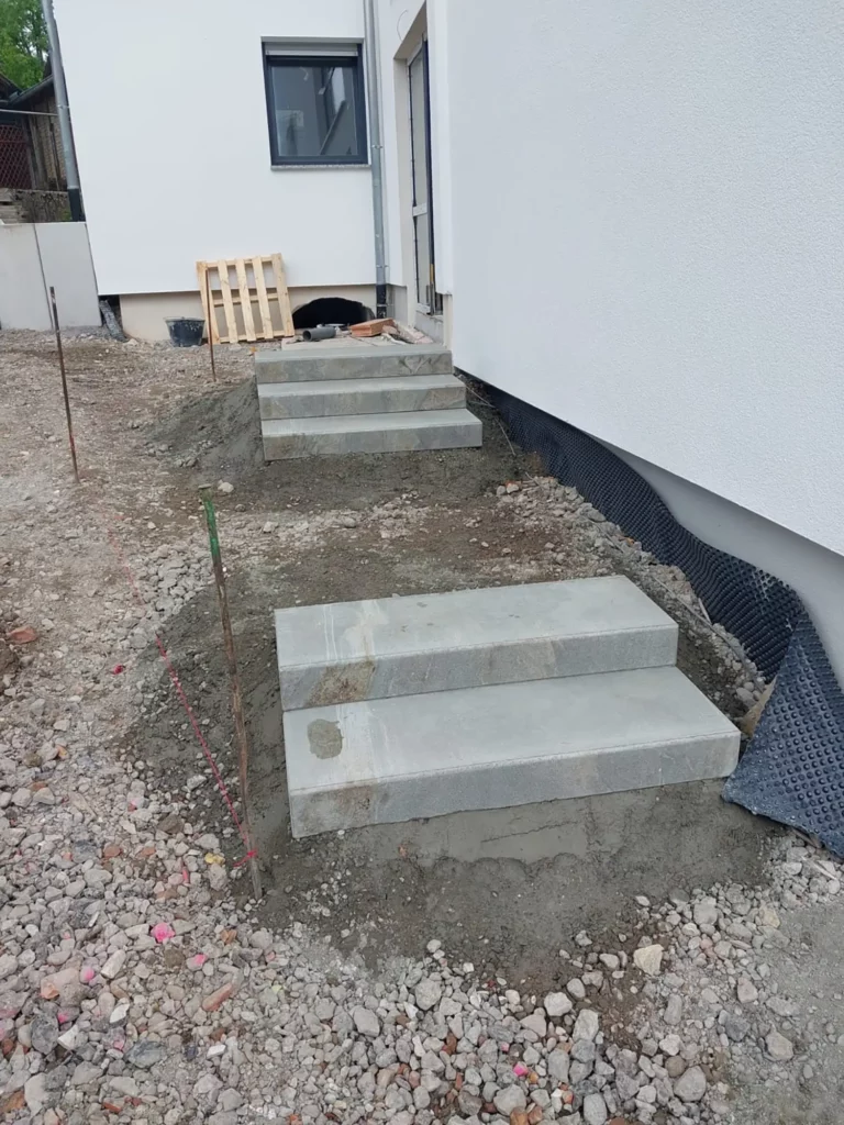 Zwei unvollendete Betonstufen neben einem weißen Gebäude auf einer Kiesfläche.